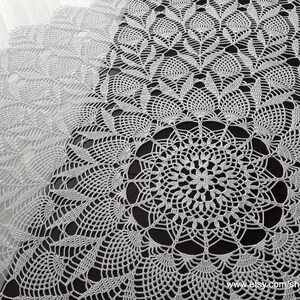 May include: A white, intricately crocheted table runner with a detailed floral and geometric pattern. The delicate lace design features a central circular motif and repeating leaf-like shapes, creating a textured surface. The runner is displayed on a dark surface.