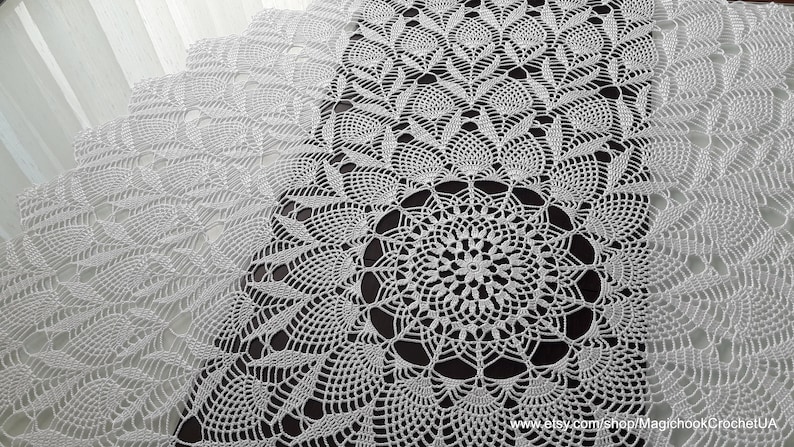 May include: A white, intricately crocheted table runner with a detailed floral and geometric pattern. The delicate lace design features a central circular motif and repeating leaf-like shapes, creating a textured surface. The runner is displayed on a dark surface.