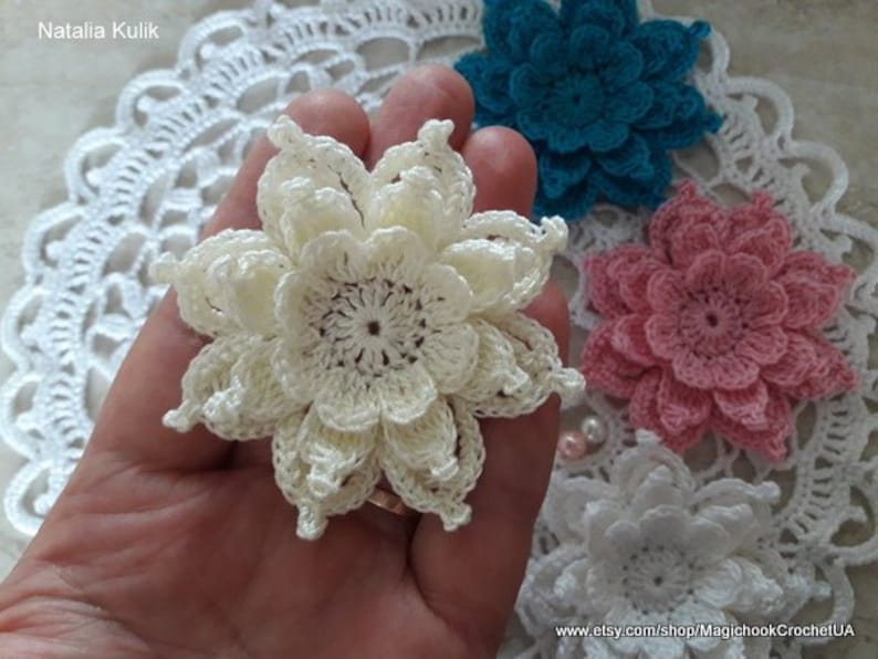May include: Three crocheted flowers in different colors: white, pink, and teal. The flowers are made with a delicate, lacy stitch and have a layered, ruffled appearance.