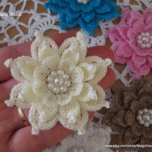May include: Five crocheted flowers with pearl centers. The flowers are in shades of white, pink, brown, and blue. The flowers are arranged on a white crocheted doily.