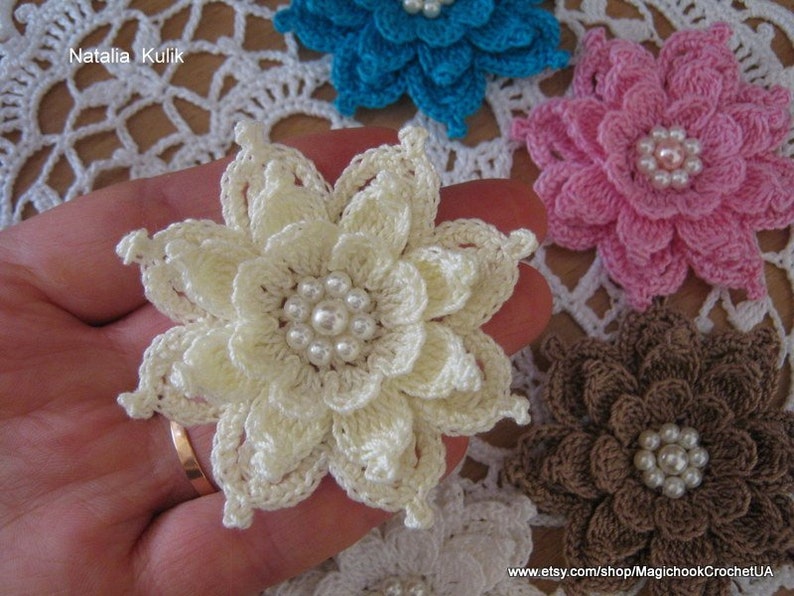 May include: Five crocheted flowers with pearl centers. The flowers are in shades of white, pink, brown, and blue. The flowers are arranged on a white crocheted doily.