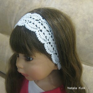 May include: A white crocheted headband with a delicate lace pattern. The headband is worn by a doll with long brown hair.