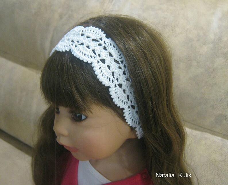 May include: A white crocheted headband with a delicate lace pattern. The headband is worn by a doll with long brown hair.