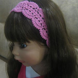 May include: A pink crocheted headband with a delicate floral pattern. The headband is worn by a doll with long brown hair.