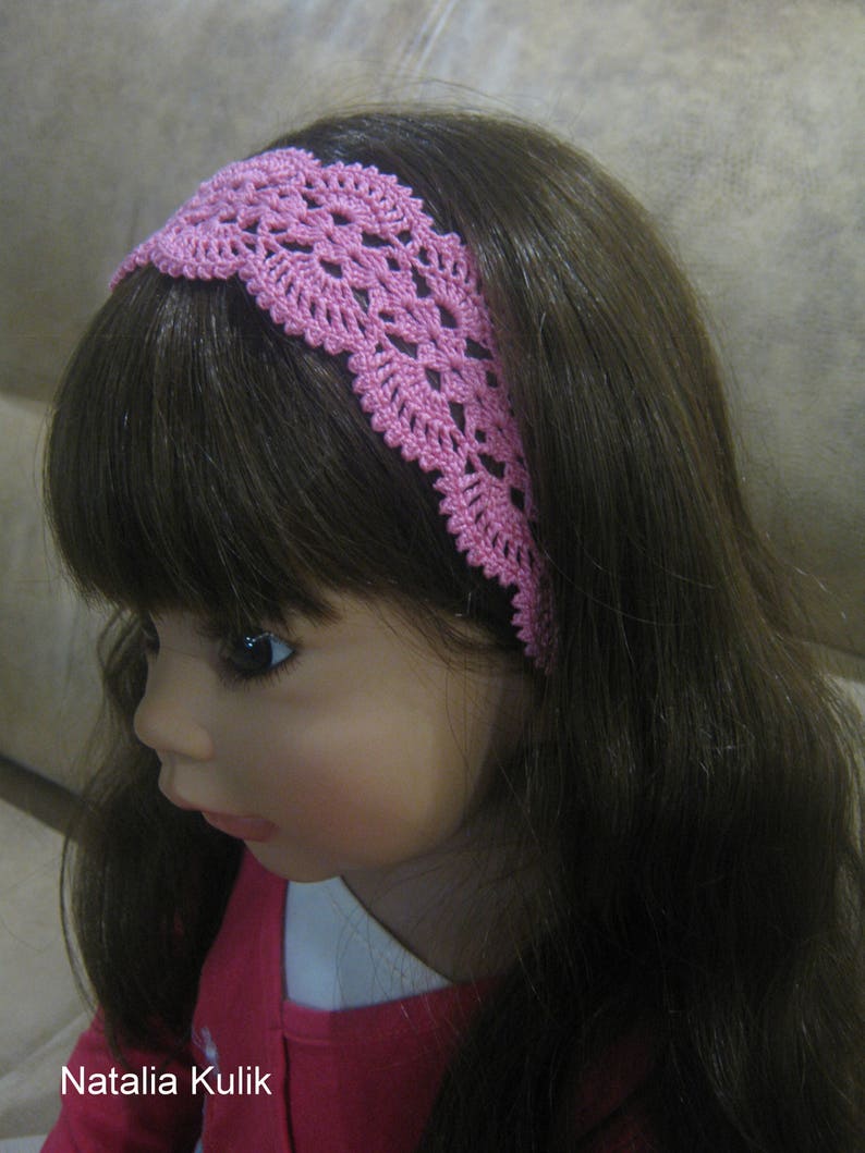May include: A pink crocheted headband with a delicate floral pattern. The headband is worn by a doll with long brown hair.