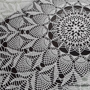 May include: White, handmade crochet doily with a detailed floral and geometric design. The doily features a central circular motif surrounded by intricate patterns. The delicate lace-like texture is visible against a dark background.