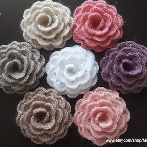 May include: Six crocheted flowers in various colors, including beige, white, pink, and purple. The flowers are arranged in a circle on a dark brown surface.