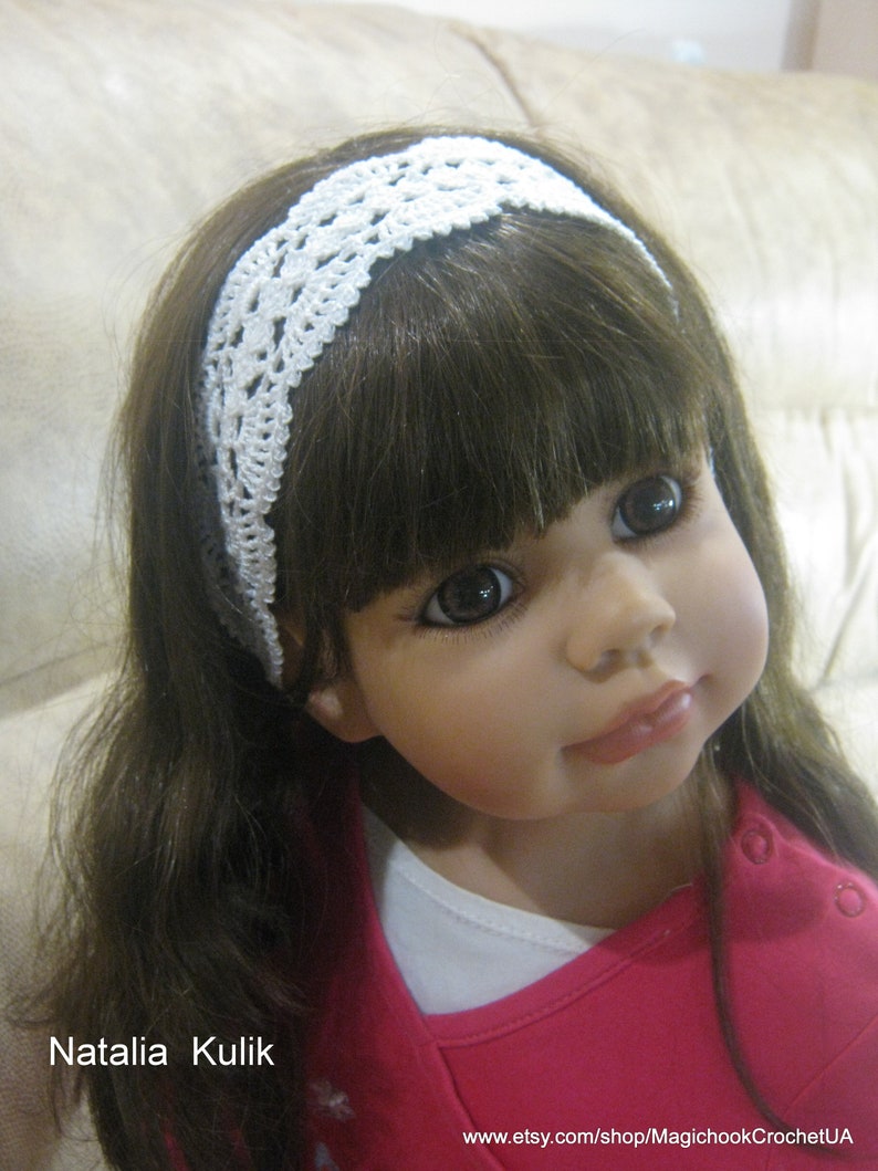 May include: A white crocheted headband with a delicate floral pattern. The headband is worn by a doll with long brown hair and a pink shirt.