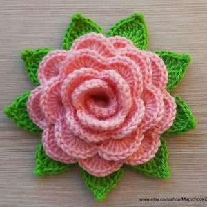 May include: A crocheted pink rose with green leaves. The rose is made of multiple layers of crocheted petals.