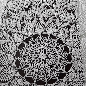 May include: White, intricately crocheted doily with a central circular design and repeating patterns. The delicate lace-like texture is visible throughout the piece, creating a detailed and elegant appearance. The doily is likely handmade.