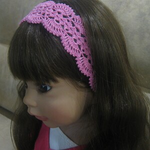 May include: A pink crocheted headband with a delicate floral pattern. The headband is worn by a doll with long brown hair.