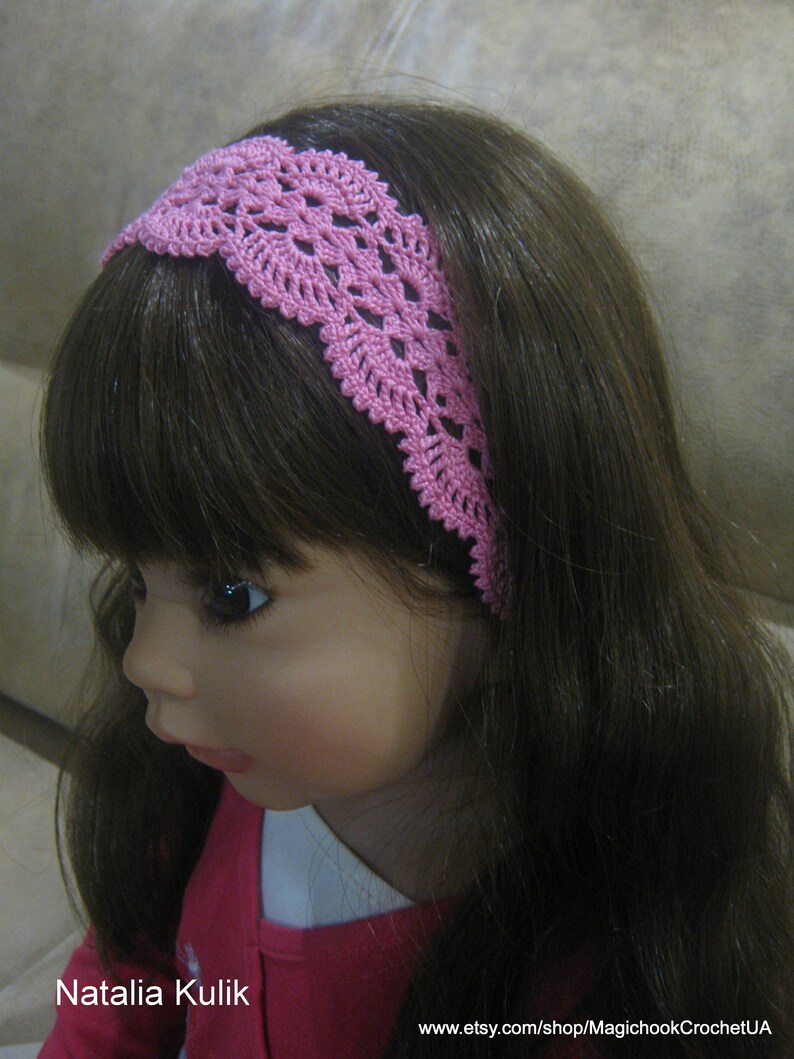 May include: A pink crocheted headband with a delicate floral pattern. The headband is worn by a doll with long brown hair.