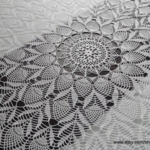 May include: A white, intricately crocheted tablecloth with a central circular design and repeating floral patterns. The delicate lace-like texture creates a visually appealing surface, perfect for a dining table or decorative display.