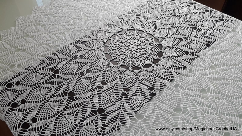 May include: A white, intricately crocheted tablecloth with a central circular design and repeating floral patterns. The delicate lace-like texture creates a visually appealing surface, perfect for a dining table or decorative display.