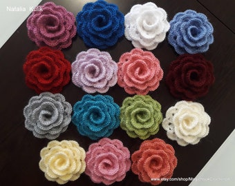 May include: A collection of 16 crocheted rose flowers in various colors, including pink, blue, red, purple, gray, teal, green, white, yellow, and orange. The flowers are arranged in a circle on a dark brown surface.