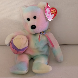 May include: A pastel-coloured Beanie Baby bear plush toy holding a small, round, pastel-coloured ball. The bear has black eyes and a black nose. A red heart-shaped tag is attached to the bear's ear.