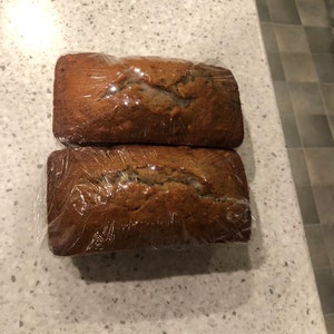 May include: Two loaves of banana bread wrapped in plastic wrap. The bread is a light brown color and has a slightly crumbly texture.