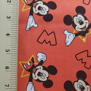Puede incluir: Tela roja con un patr&oacute;n repetido de Mickey Mouse. Mickey lleva pantalones cortos rojos y zapatos amarillos. Est&aacute; sosteniendo una estrella amarilla con un centro rojo. El fondo es de color rojo s&oacute;lido.