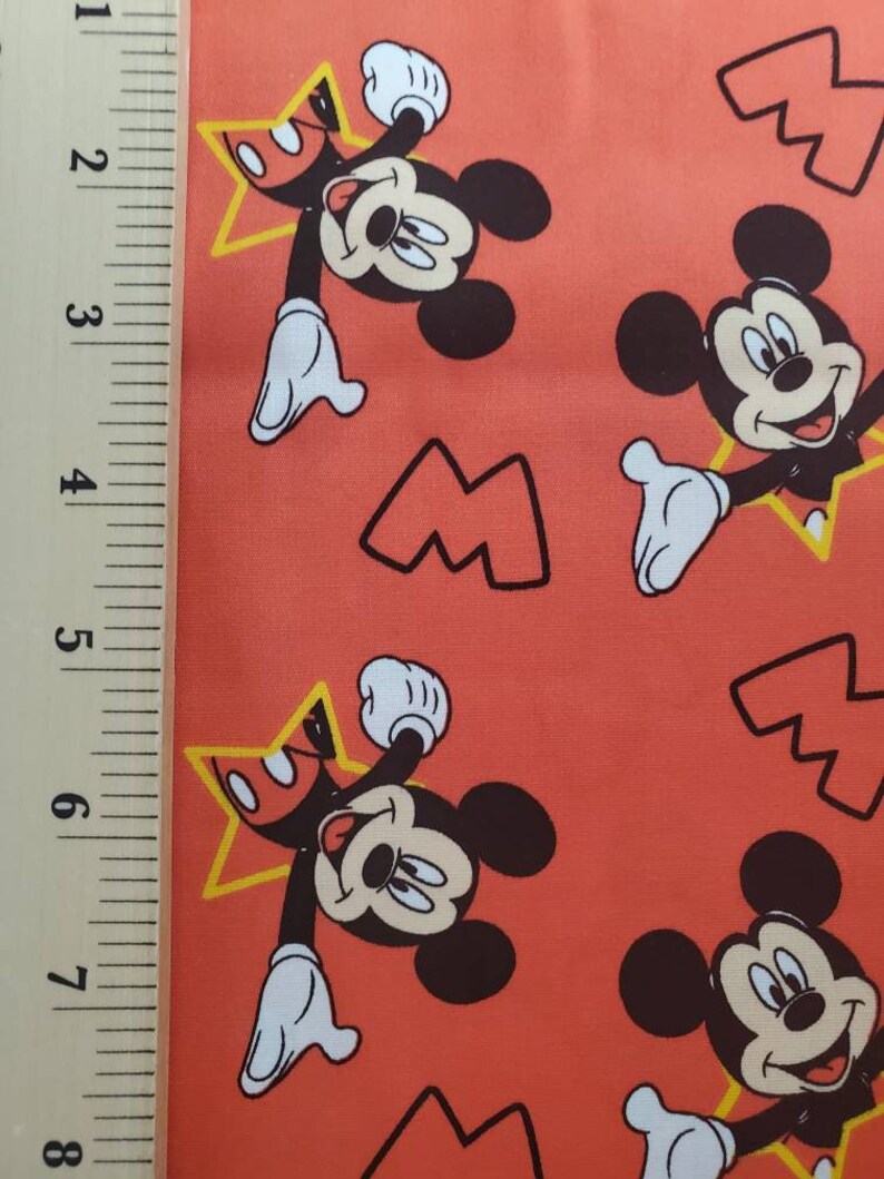 Puede incluir: Tela roja con un patr&oacute;n repetido de Mickey Mouse. Mickey lleva pantalones cortos rojos y zapatos amarillos. Est&aacute; sosteniendo una estrella amarilla con un centro rojo. El fondo es de color rojo s&oacute;lido.