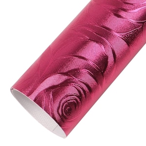 May include: A roll of metallic fuchsia faux leather fabric featuring a raised, embossed rose pattern across the surface. The material has a shiny, reflective finish with a white backing visible at the edge of the rolled sheet of craft material.