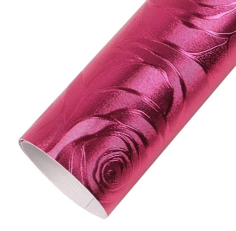 May include: A roll of metallic fuchsia faux leather fabric featuring a raised, embossed rose pattern across the surface. The material has a shiny, reflective finish with a white backing visible at the edge of the rolled sheet of craft material.