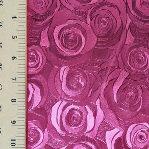 May include: A vibrant magenta fabric featuring a repeating embossed rose pattern with a glossy finish. A wooden ruler is positioned along the left edge, showing a measurement scale of 12 inches to provide a clear sense of the fabric's scale.
