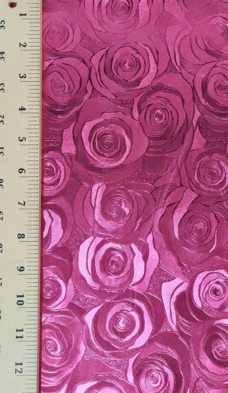 May include: A vibrant magenta fabric featuring a repeating embossed rose pattern with a glossy finish. A wooden ruler is positioned along the left edge, showing a measurement scale of 12 inches to provide a clear sense of the fabric's scale.
