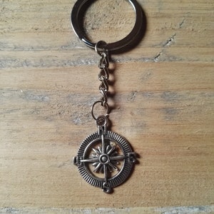 Puede incluir: Llavero de brújula de plata con una cadena y un anillo. La brújula tiene una estrella de plata en el centro y las letras N, S, E y W alrededor del borde.