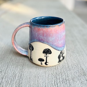 May include: A ceramic mug with a colorful glaze, featuring a gradient of pink, purple, and blue. The mug has a cream-colored base with black and white mushroom illustrations. The handle and rim are also colored.