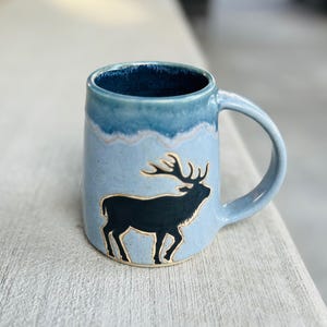 Könnte beinhalten: Hellblaue Keramiktasse mit dunkelblauem Innenraum. Die Tasse zeigt eine schwarze Silhouette eines Hirsches mit Geweih. Henkel und Rand sind ebenfalls blau. Ein einzigartiges Trinkgefäß.