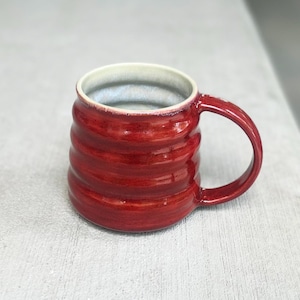 May include: A glossy, deep red ceramic mug featuring a series of horizontal, rounded ridges along its exterior body. The mug has a curved handle on the right side and a smooth, light-colored interior finish that contrasts with the vibrant red glaze.