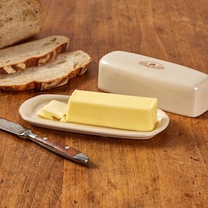 May include: A rectangular cream-colored ceramic butter dish with a matching lid featuring a small brown acorn graphic. A solid block of yellow butter sits on the base with a few slices cut off, accompanied by a metal knife with a wooden handle.