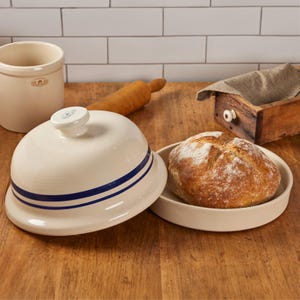 May include: A cream-colored ceramic bread cloche with a matching base and a domed lid featuring two horizontal navy blue stripes. The lid has a small knob handle on top. A round, crusty loaf of bread sits inside the open base of the cloche.