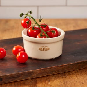 May include: A cream-colored ceramic ramekin featuring the brown text Acorn Bluff and an acorn logo. The ramekin is filled with a cluster of bright red cherry tomatoes on a green vine, with two additional loose cherry tomatoes placed nearby.