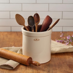 May include: A cream-colored ceramic crock featuring the text Acorn Stamp with a small acorn graphic. The crock holds five wooden kitchen utensils of varying shades and shapes. A wooden rolling pin rests on a tan cloth in front of the crock.
