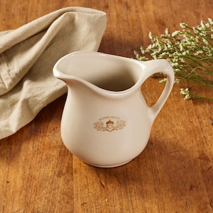 May include: A cream-colored ceramic pitcher with a curved handle and a pouring spout. The front of the pitcher features a brown printed logo with an acorn icon, oak leaves, and the text Acorn Bluff written in a small, clear font.