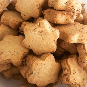 May include: A close-up shot of a pile of star-shaped cookies. The cookies are golden brown with a slightly textured surface and appear to have small, dark specks throughout. The cookies are stacked and overlapping, filling the frame.