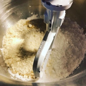 K&ouml;nnte beinhalten: Nahaufnahme eines silbernen Standmixers mit einem wei&szlig;en Paddel-Aufsatz. Die Sch&uuml;ssel ist mit einer Mischung aus wei&szlig;en und hellbraunen Zutaten gef&uuml;llt.