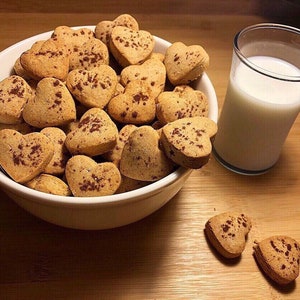 May include: A white bowl filled with heart-shaped cookies, some of which have fallen out of the bowl. The cookies are a light brown color and have a sprinkle of cinnamon on top. A glass of milk is next to the bowl.