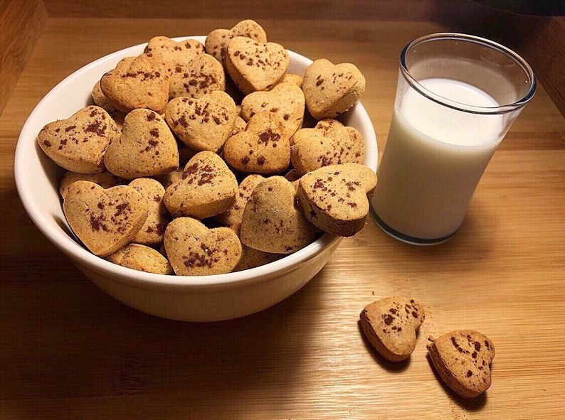 May include: A white bowl filled with heart-shaped cookies, some of which have fallen out of the bowl. The cookies are a light brown color and have a sprinkle of cinnamon on top. A glass of milk is next to the bowl.