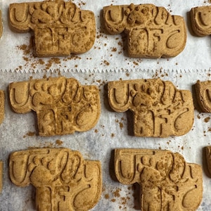 K&ouml;nnte beinhalten: Nahaufnahme einer Backform mit mehreren braunen Hundekekse. Die Kekse sind in Form eines Hundebeins geformt und haben die Worte "US ALL" und "AFC" eingepr&auml;gt. Die Kekse sind mit Zimt best&auml;ubt.