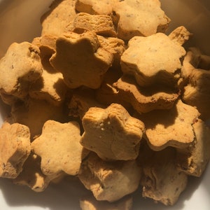 May include: A close-up shot of a white bowl filled with star-shaped cookies. The cookies are golden brown with a slightly textured surface. The cookies appear to be homemade and are stacked on top of each other.