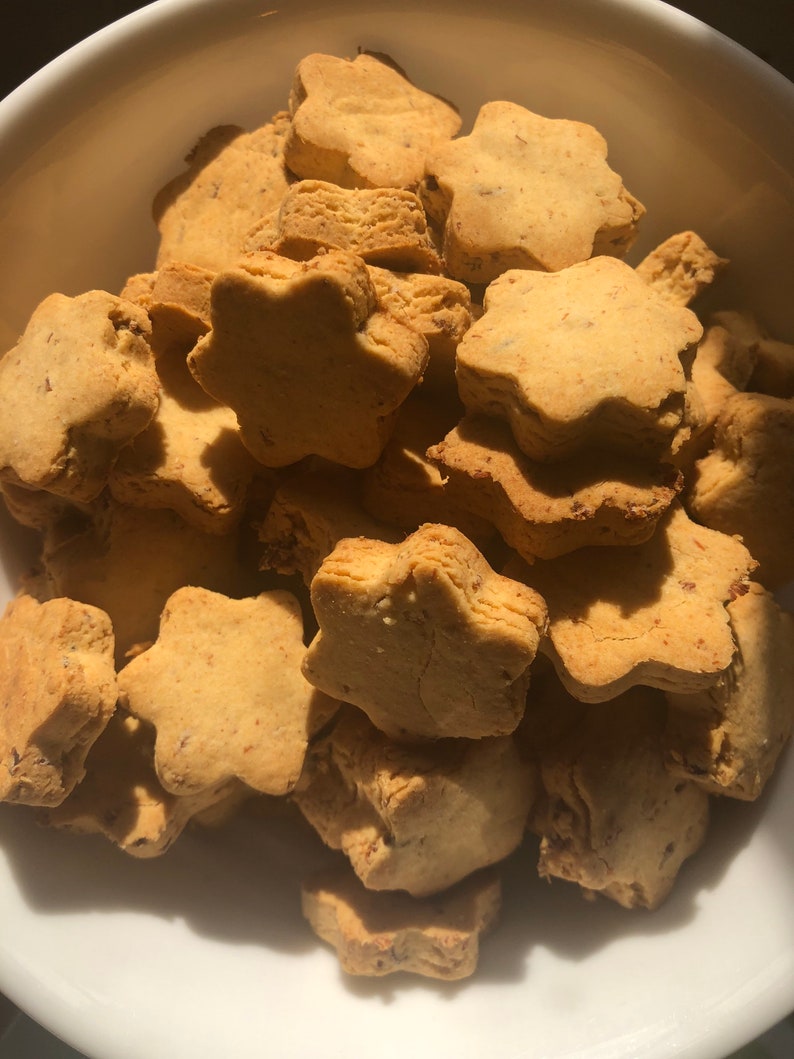 May include: A close-up shot of a white bowl filled with star-shaped cookies. The cookies are golden brown with a slightly textured surface. The cookies appear to be homemade and are stacked on top of each other.