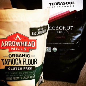 May include: Two bags of gluten-free flour. A white bag with a red logo that says "Arrowhead Mills" and "Organic Tapioca Flour". A black bag with a white logo that says "Terrasoul Superfoods" and "Coconut Flour".