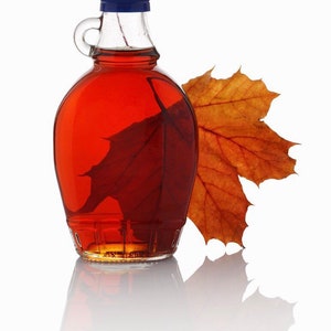 May include: A clear glass maple syrup bottle with a blue cap, filled with amber syrup. A maple leaf is inside the bottle, and another leaf rests beside it. The bottle is reflected on a white surface.
