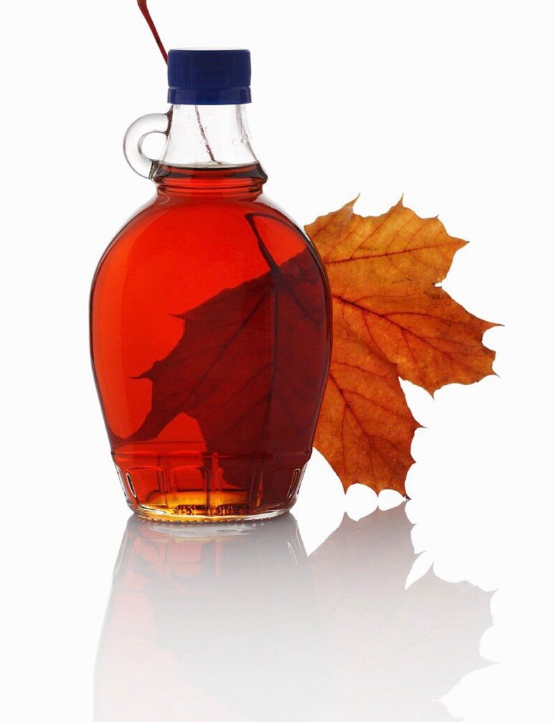 May include: A clear glass maple syrup bottle with a blue cap, filled with amber syrup. A maple leaf is inside the bottle, and another leaf rests beside it. The bottle is reflected on a white surface.
