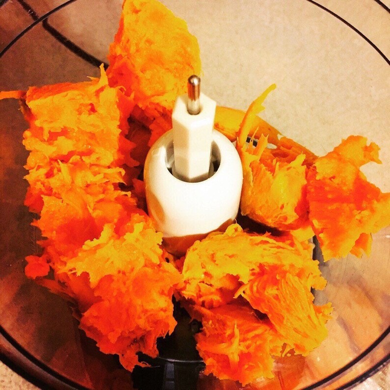 May include: A close-up of a food processor bowl filled with shredded orange sweet potatoes. The food processor blade is visible in the center of the bowl.