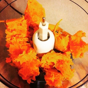May include: A close-up shot of a food processor bowl filled with bright orange, shredded pumpkin. The white central component of the processor is visible, surrounded by the pumpkin. The image is well-lit, highlighting the texture of the pumpkin.