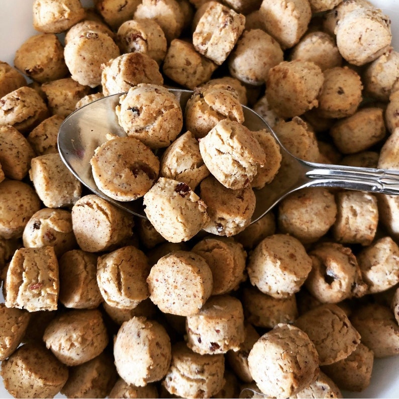 May include: A close-up image of a bowl filled with small, round, light brown cereal pieces. A silver spoon is partially submerged in the cereal, with some pieces clinging to the spoon.