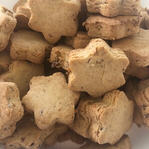 May include: A close-up shot of a pile of star-shaped cookies. The cookies are a light brown color with a slightly rough texture. Some cookies have visible dark specks, possibly from added ingredients. The cookies are stacked together, filling the frame.